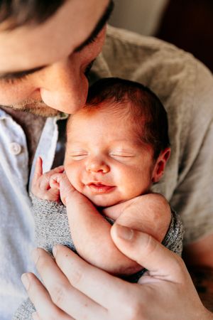 Father kissing his smiling newborn baby held close to his chest in warm light.