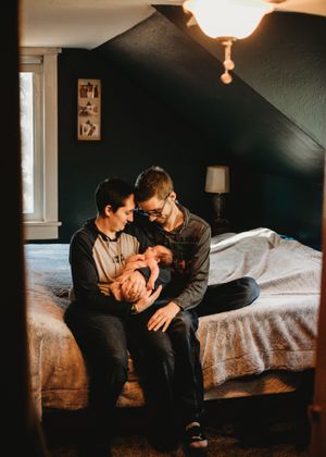 Two parents sitting on bed in dark room lovingly holding newborn baby.
