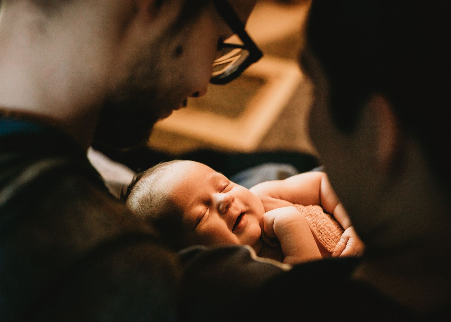 Two parents gazing at sleeping newborn baby held between them closely.
