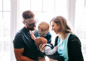 A joyful family of four with parents holding a newborn and toddler near a bright window.