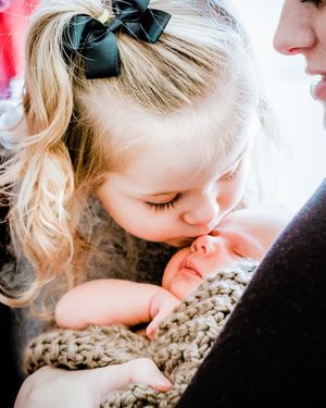 Young blonde girl with bow kissing newborn sibling wrapped in knit blanket.