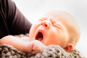 Newborn baby yawning while being cradled in a parent's arms on knit blanket.