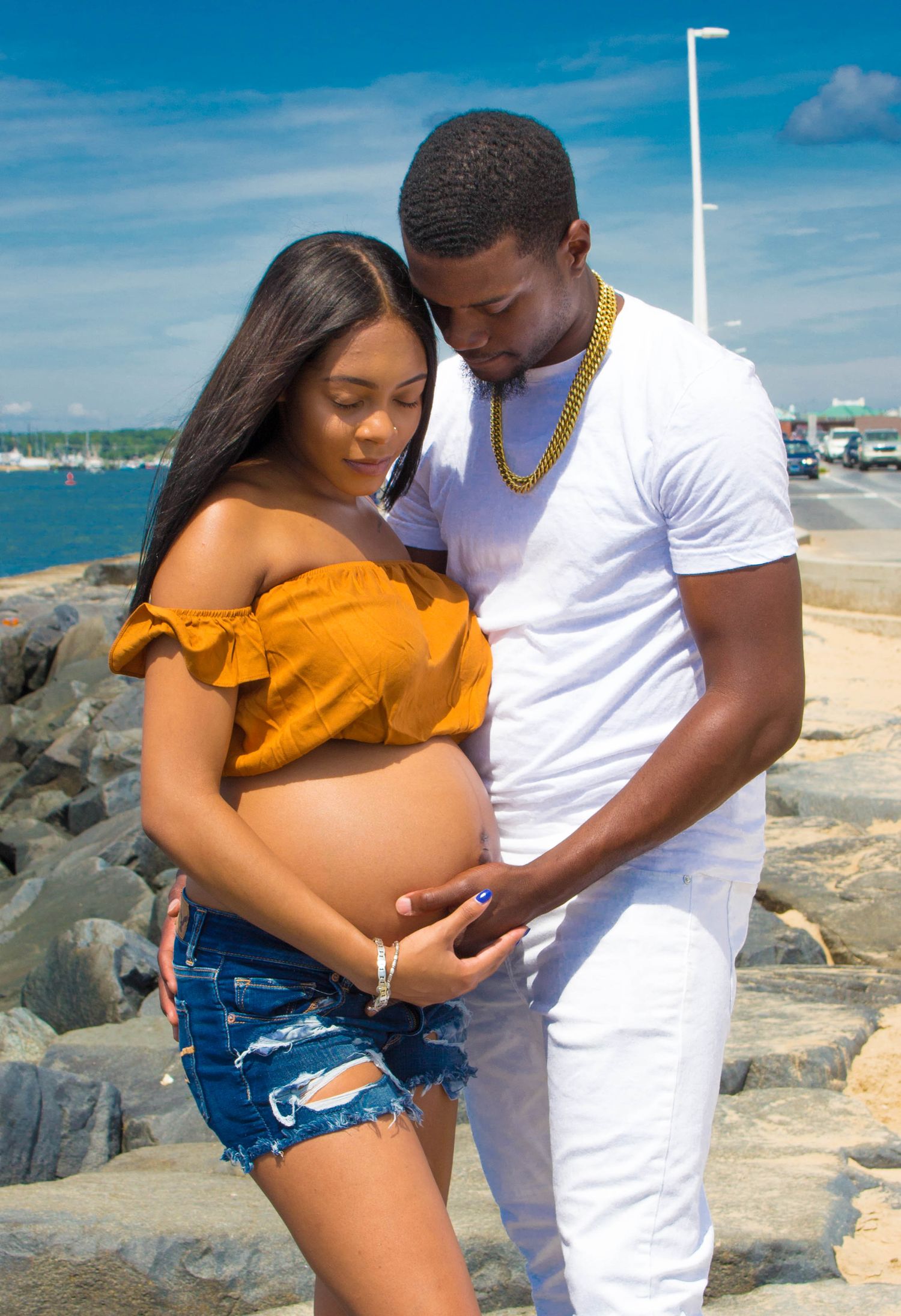 Expecting couple in casual summer outfits embrace on rocky beach. Maternity photoshoot. L. Visions Photography.