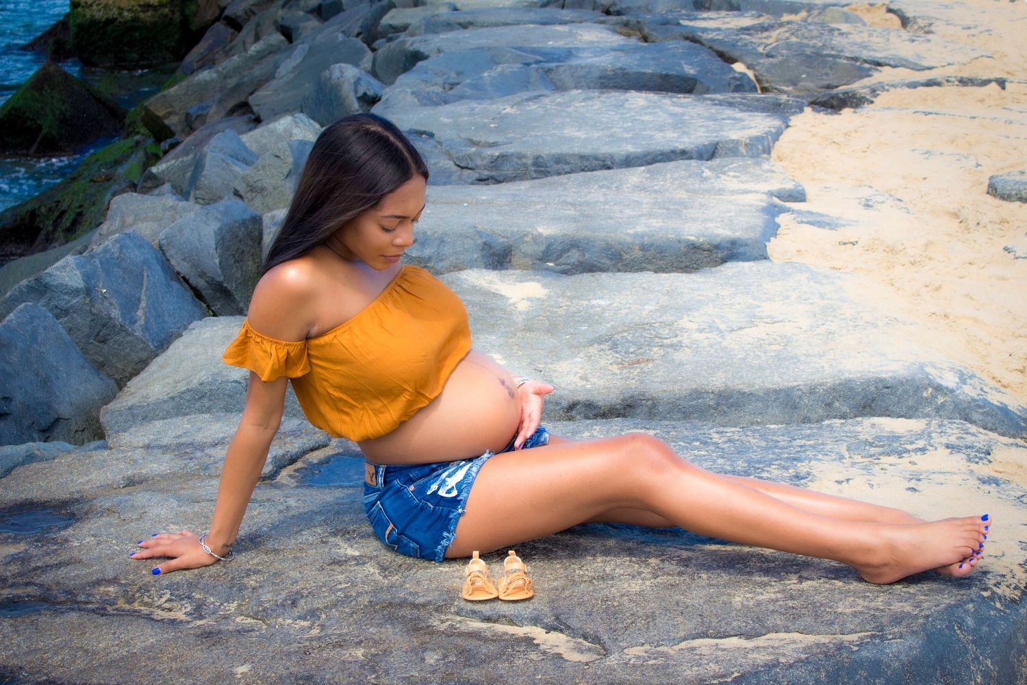 Mother, Woman in yellow top and denim shorts poses on rocky shoreline. Maternity photoshoot. L. Visions Photography.