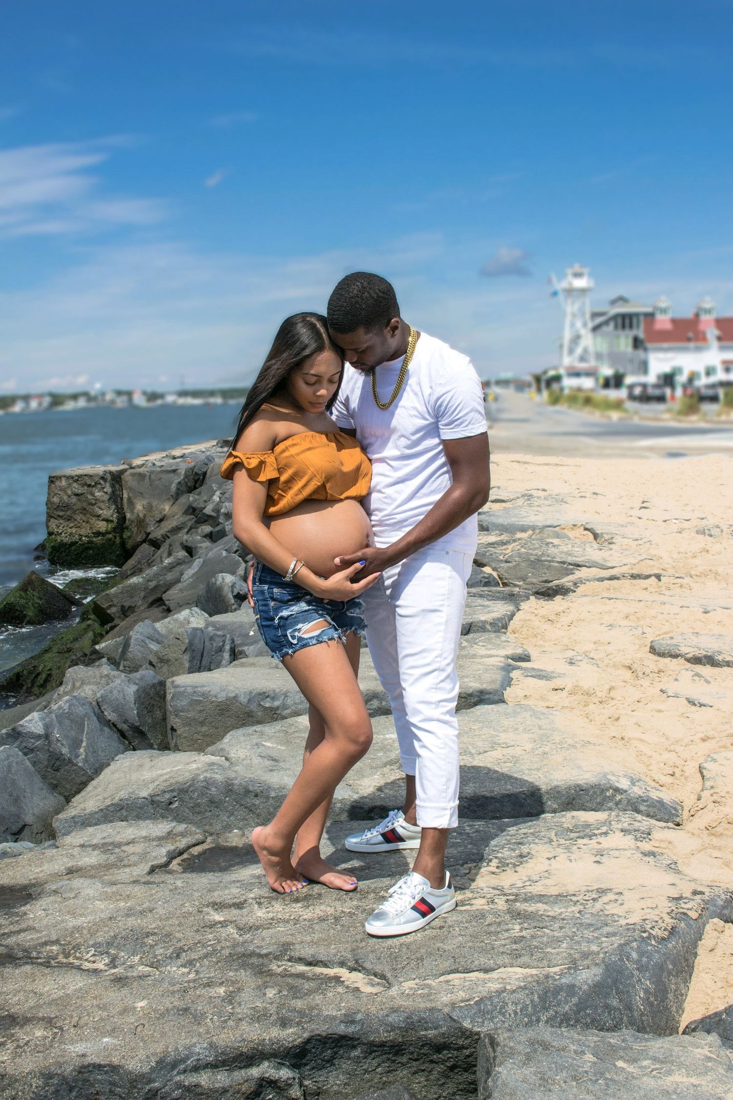 A couple shares intimate moments on rocky beach terrain with coastal homes in background. Maternity photoshoot. L. Visions Photography