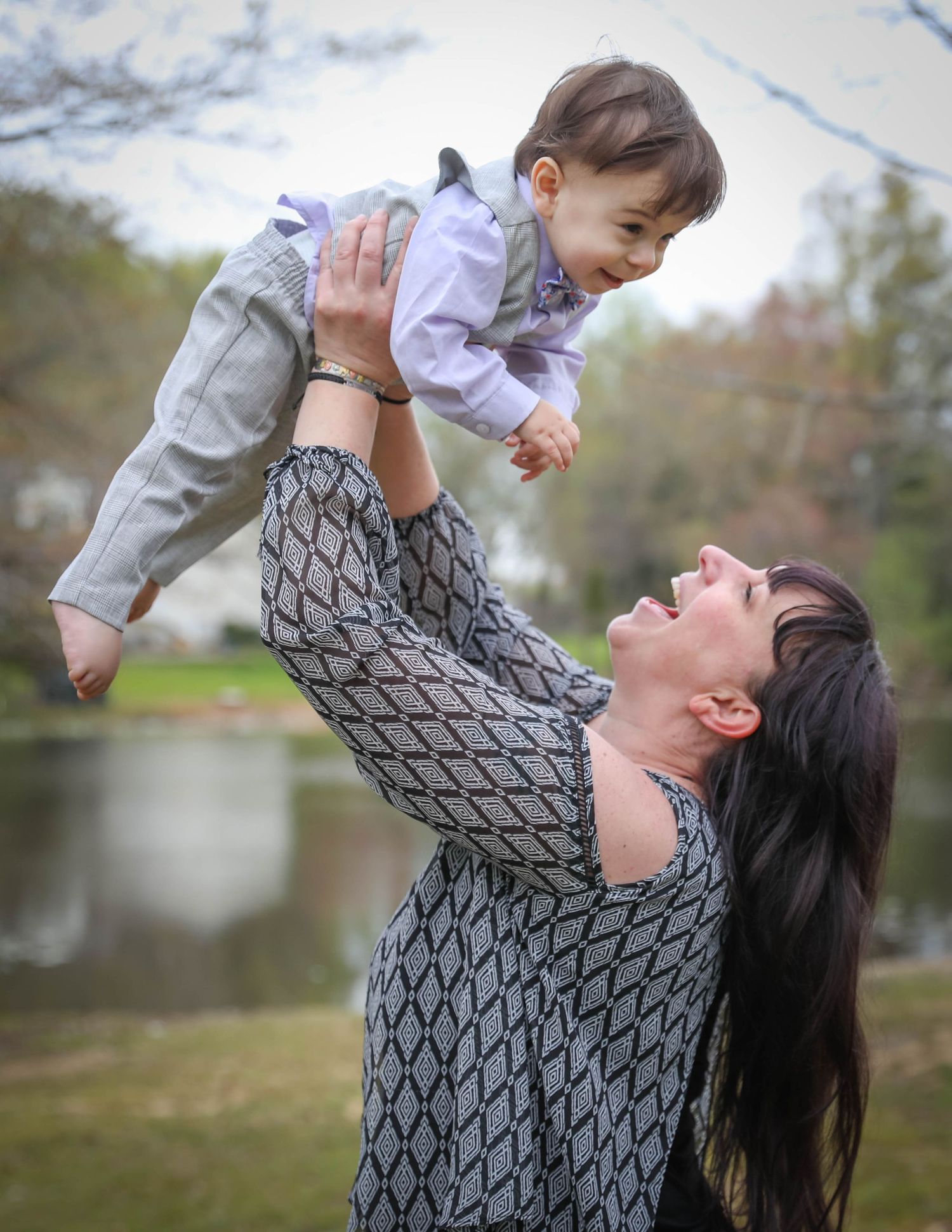 Playful parent lifts smiling baby high in the air during outdoor photoshoot. Family photoshoot. L. Visions Photography. Mother and son.