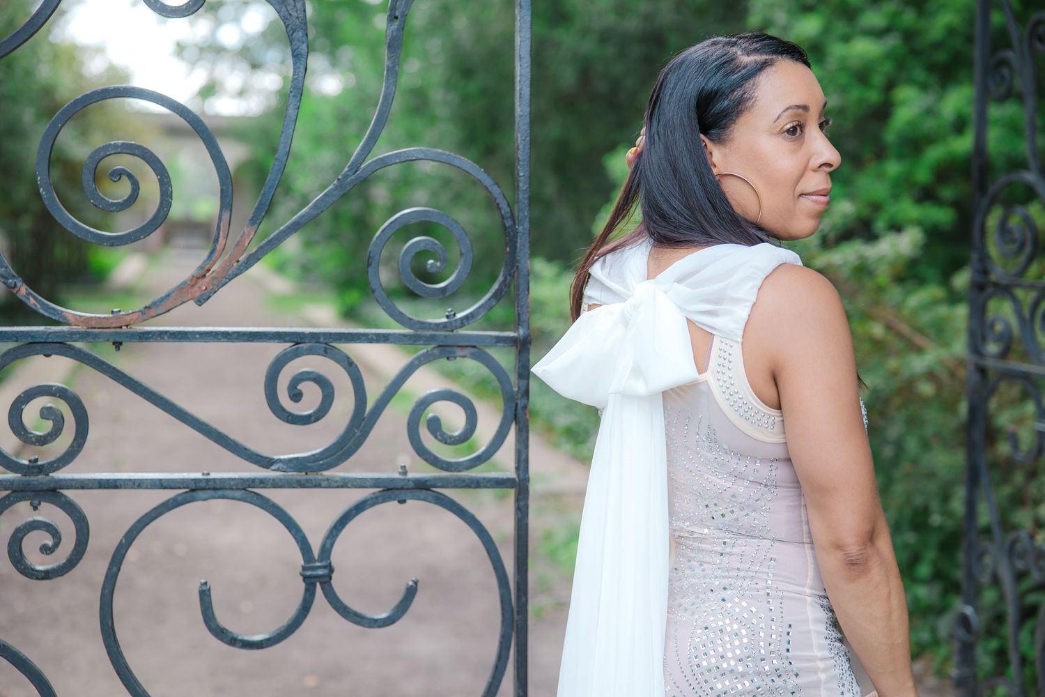 Woman in white dress poses against decorative iron gate in garden setting. Lifestyle photoshoot. L. Visions Photography