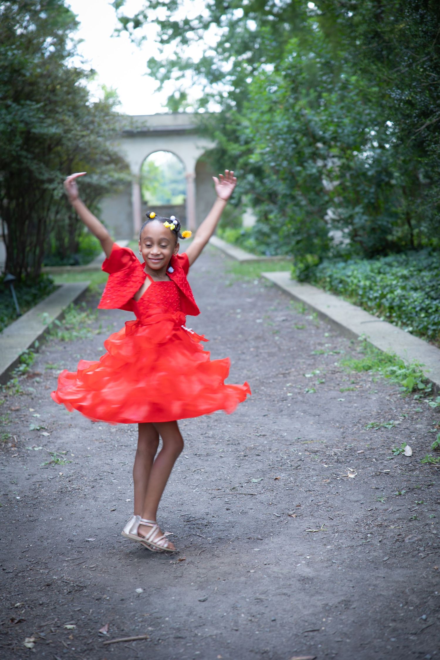 Young dancer in flowing red dress twirls joyfully on garden path near stone archway. Lifestyle photoshoot. L. Visions Photography