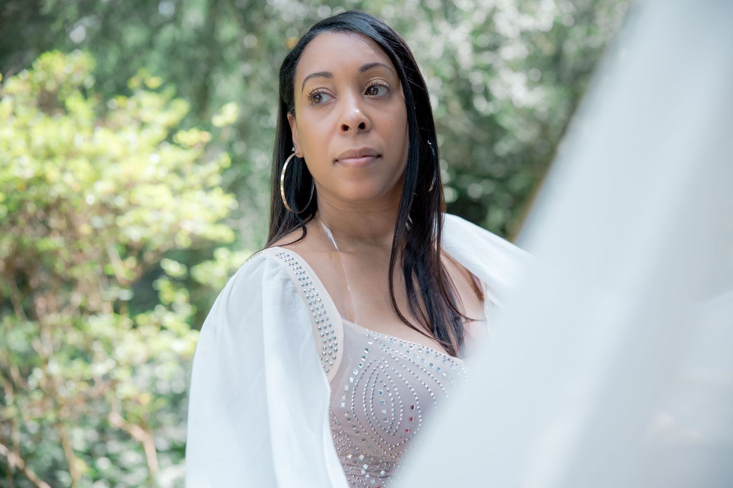 Woman wearing white lace dress stands elegantly in an outdoor garden setting. Lifestyle photoshoot. L. Visions Photography