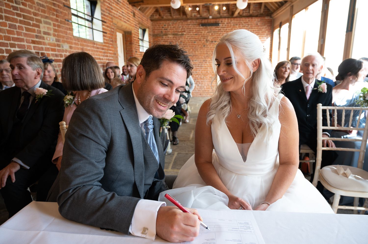 Couple signs marriage documents at table in rustic brick venue during wedding ceremony.