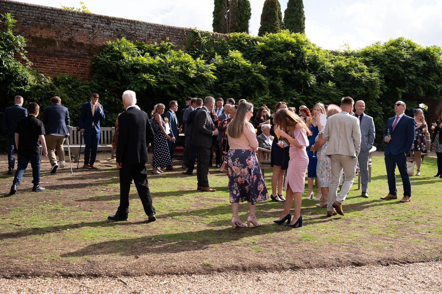 Wedding guests mingling on lawn with historic brick wall and cypress trees in background.