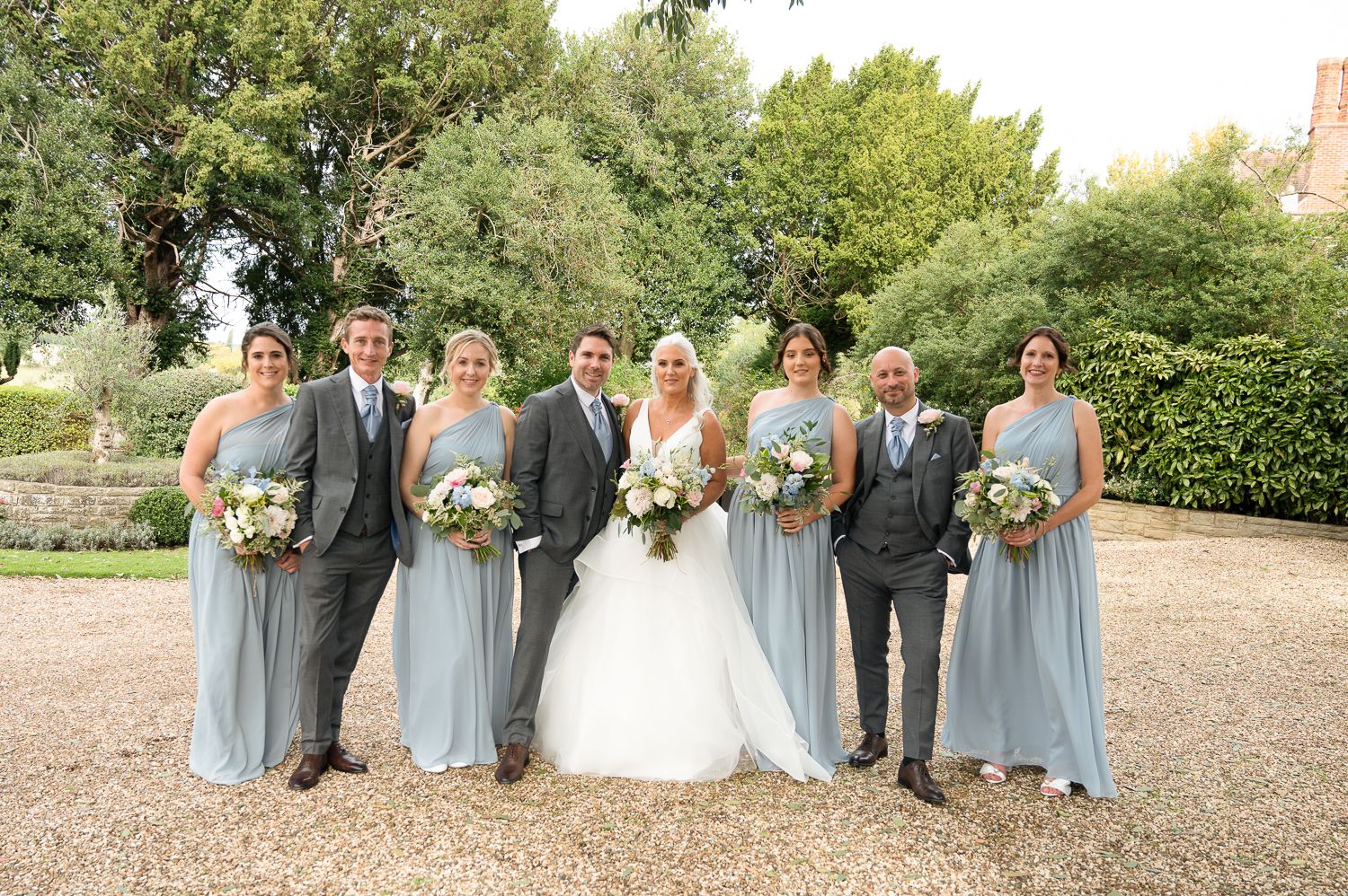 Wedding party poses for formal photo in pale blue attire with garden backdrop.