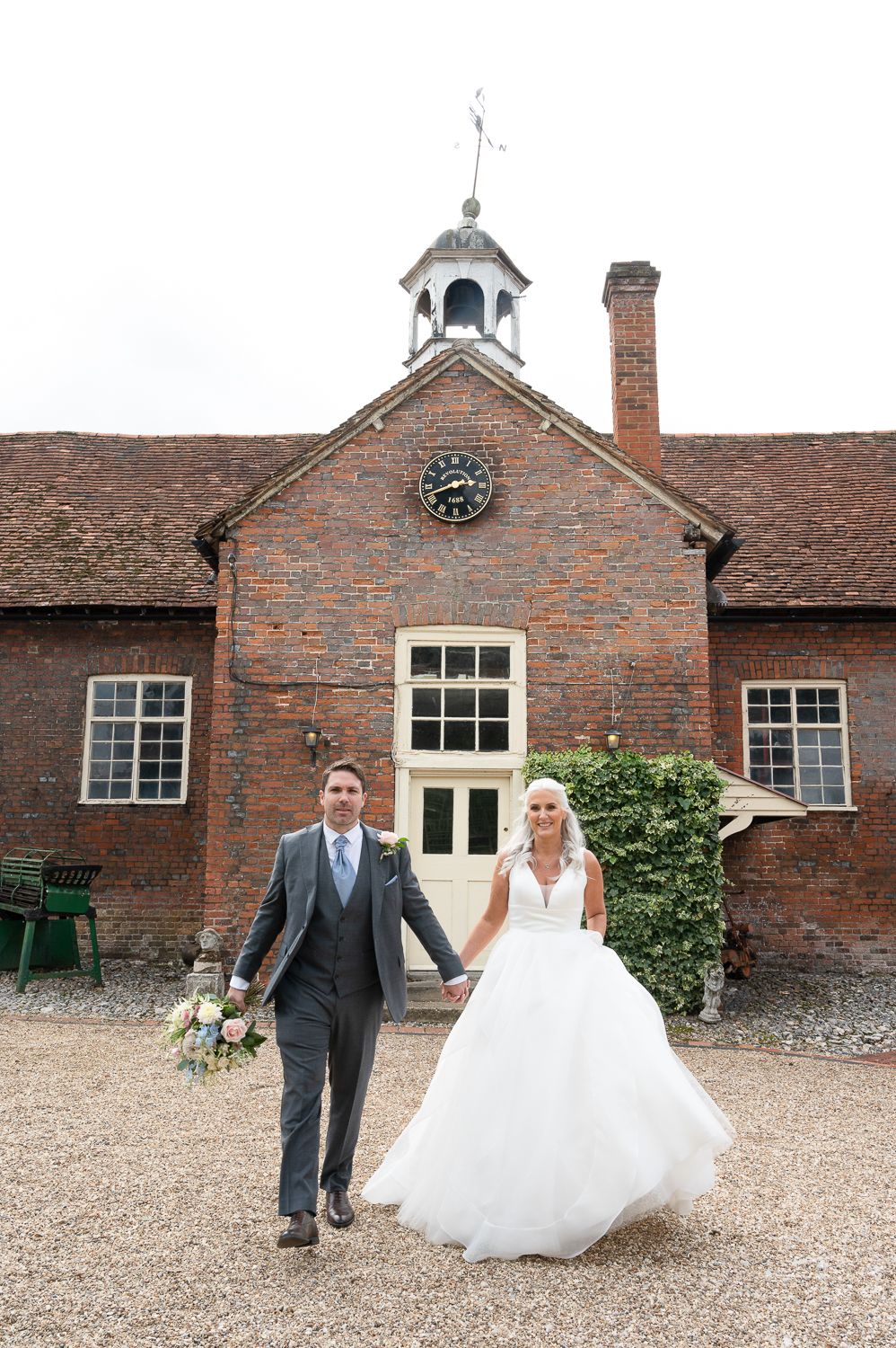 Newlyweds hold hands walking outside historic brick building with clock tower and white trim.