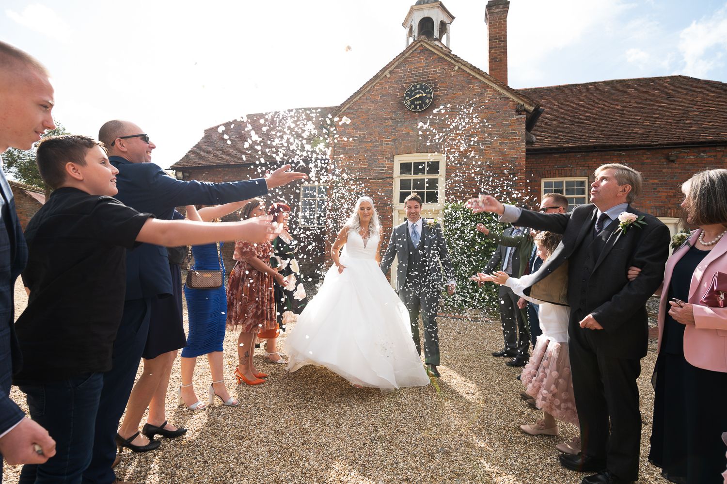 Newlyweds walk through confetti shower thrown by wedding guests at countryside venue.