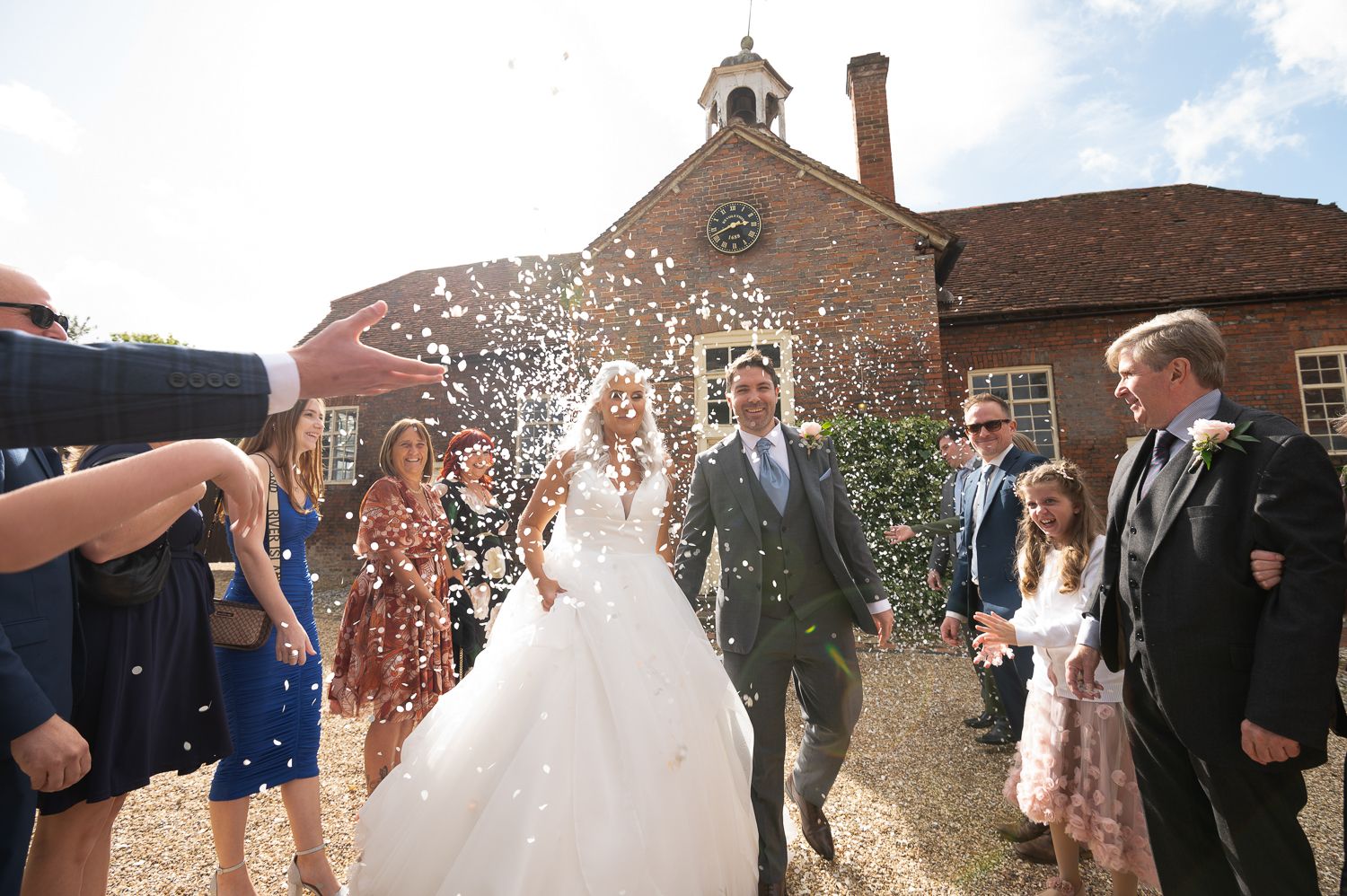 Wedding couple walks through confetti shower outside historic brick building with guests celebrating.