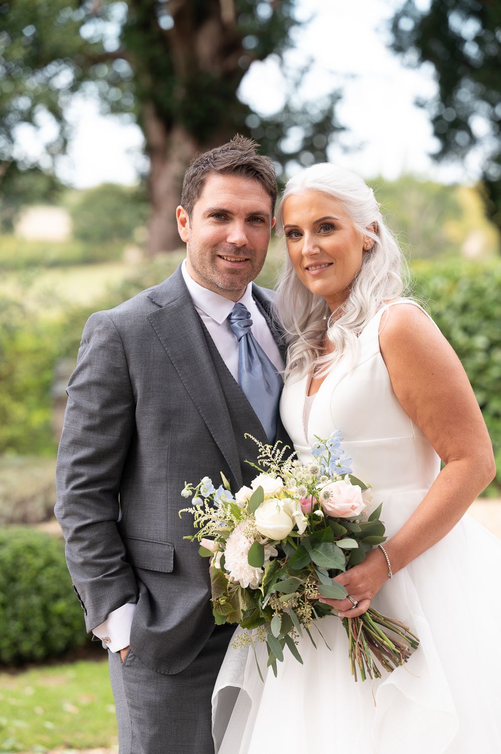 Wedding couple poses for portrait with elegant white floral bouquet in garden setting.
