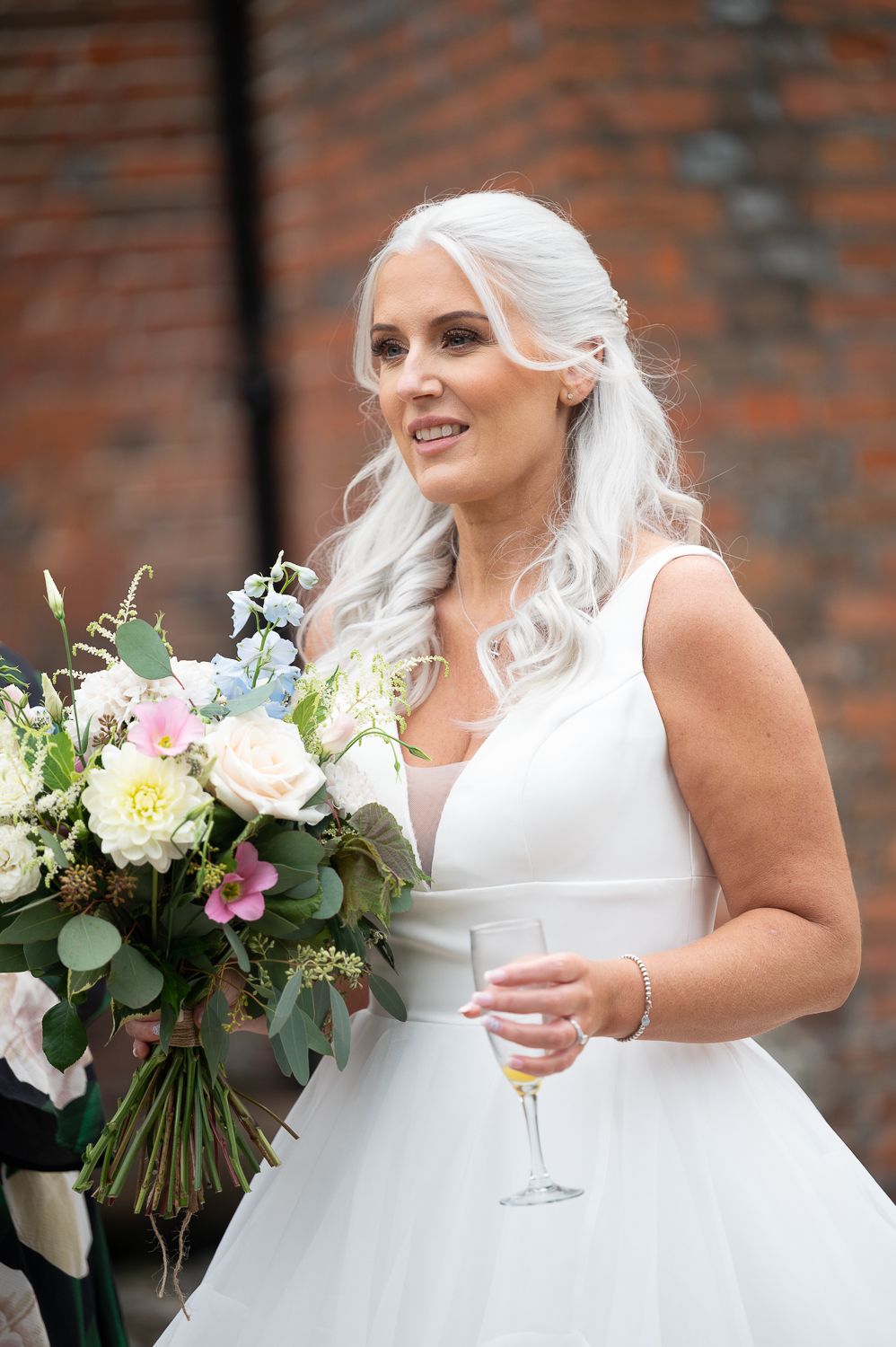 Bride holds champagne and bouquet featuring white roses and greenery against brick backdrop.