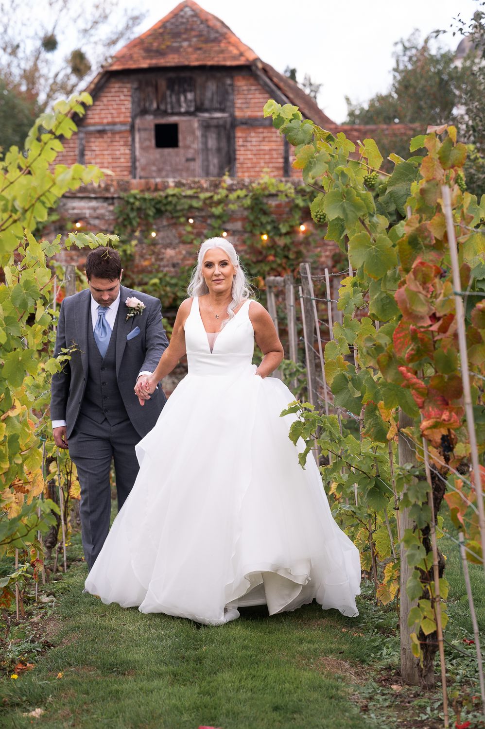 A couple walks hand-in-hand through vineyard rows with rustic brick building in background.