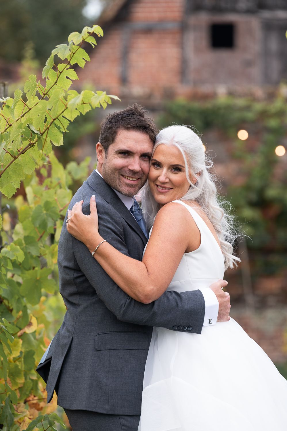 Happy couple embrace in garden setting with rustic brick building visible behind them.