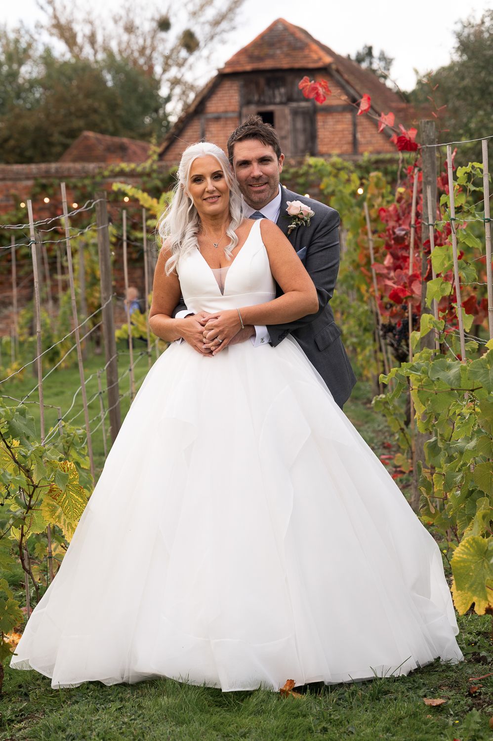 Wedding couple poses in front of rustic brick building surrounded by autumn vineyard foliage.