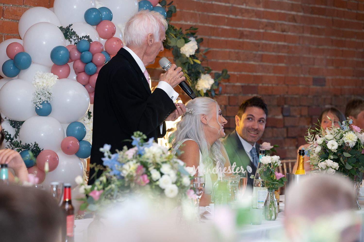 Wedding reception with balloon decor and floral centerpieces at head table with brick wall backdrop.