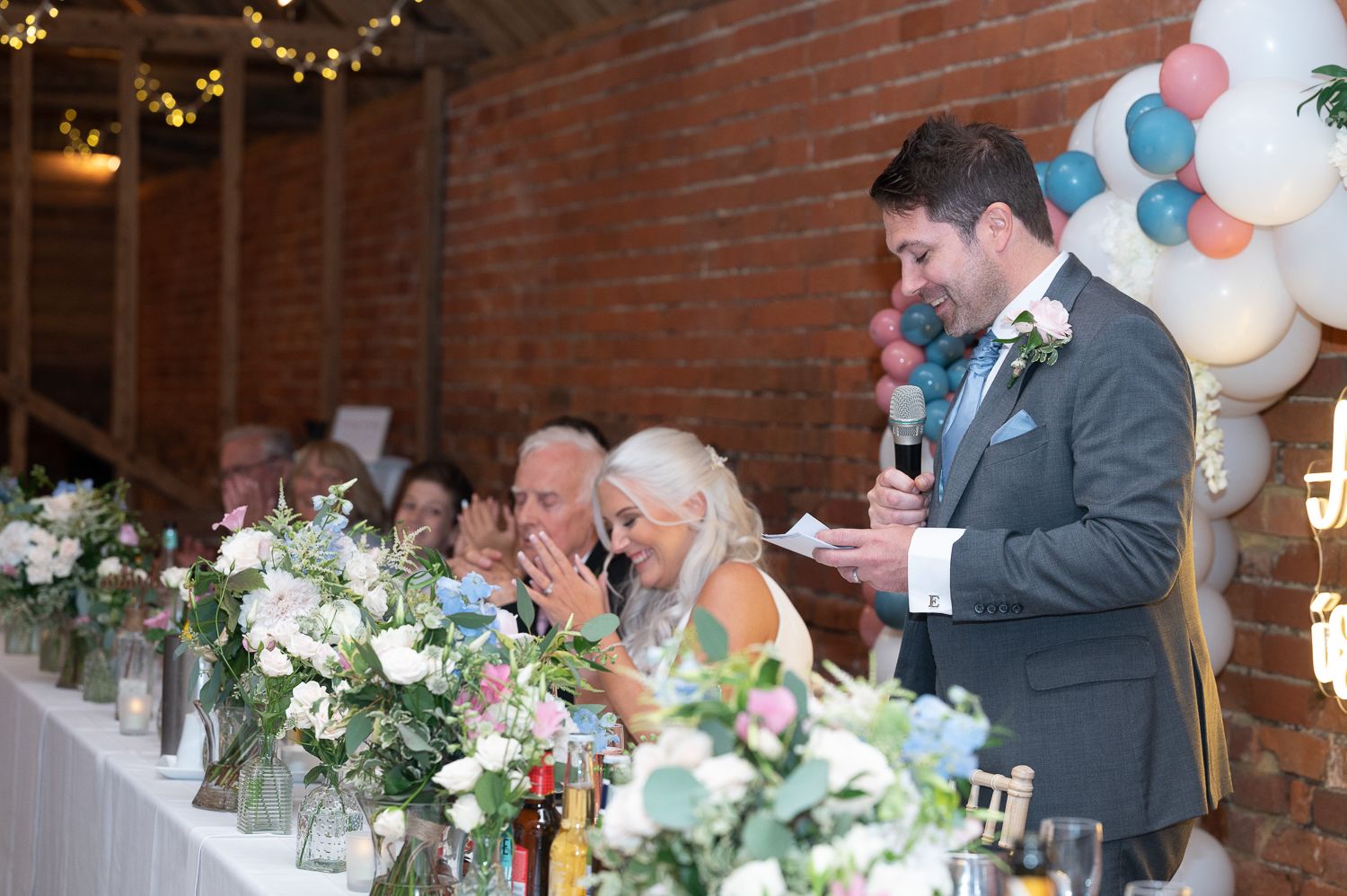 Wedding reception speeches at decorated head table with white floral arrangements and brick wall background.