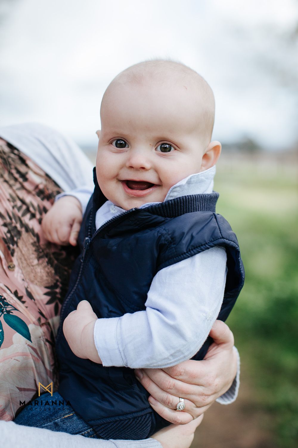 Smiling baby in navy vest and gray long-sleeve shirt being held outdoors with green fields in background.