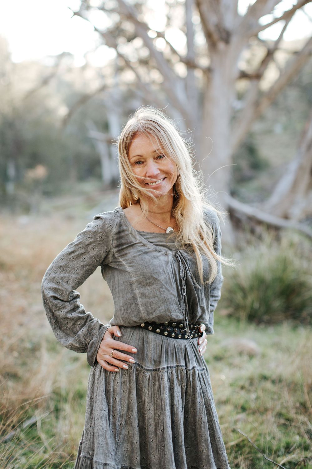 Smiling blonde woman in gray boho dress with belt posing outdoors in natural bushland setting.