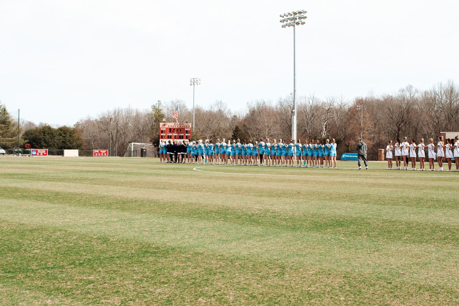 Two sports teams lined up on a grass field during pre-game ceremonies with stadium lights in the background.