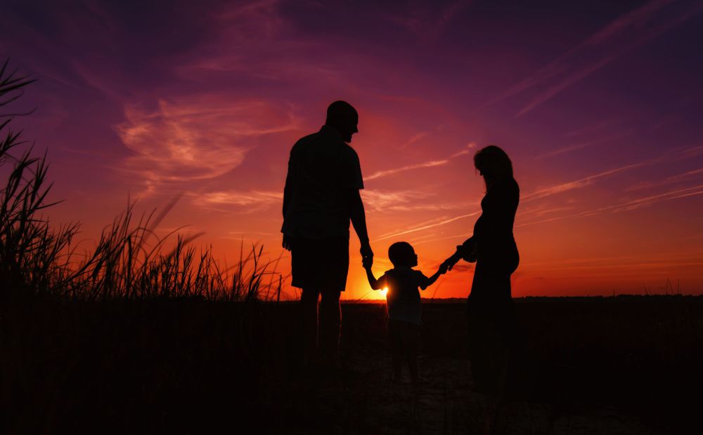 Family silhouettes hold hands against dramatic purple and orange sunset sky.