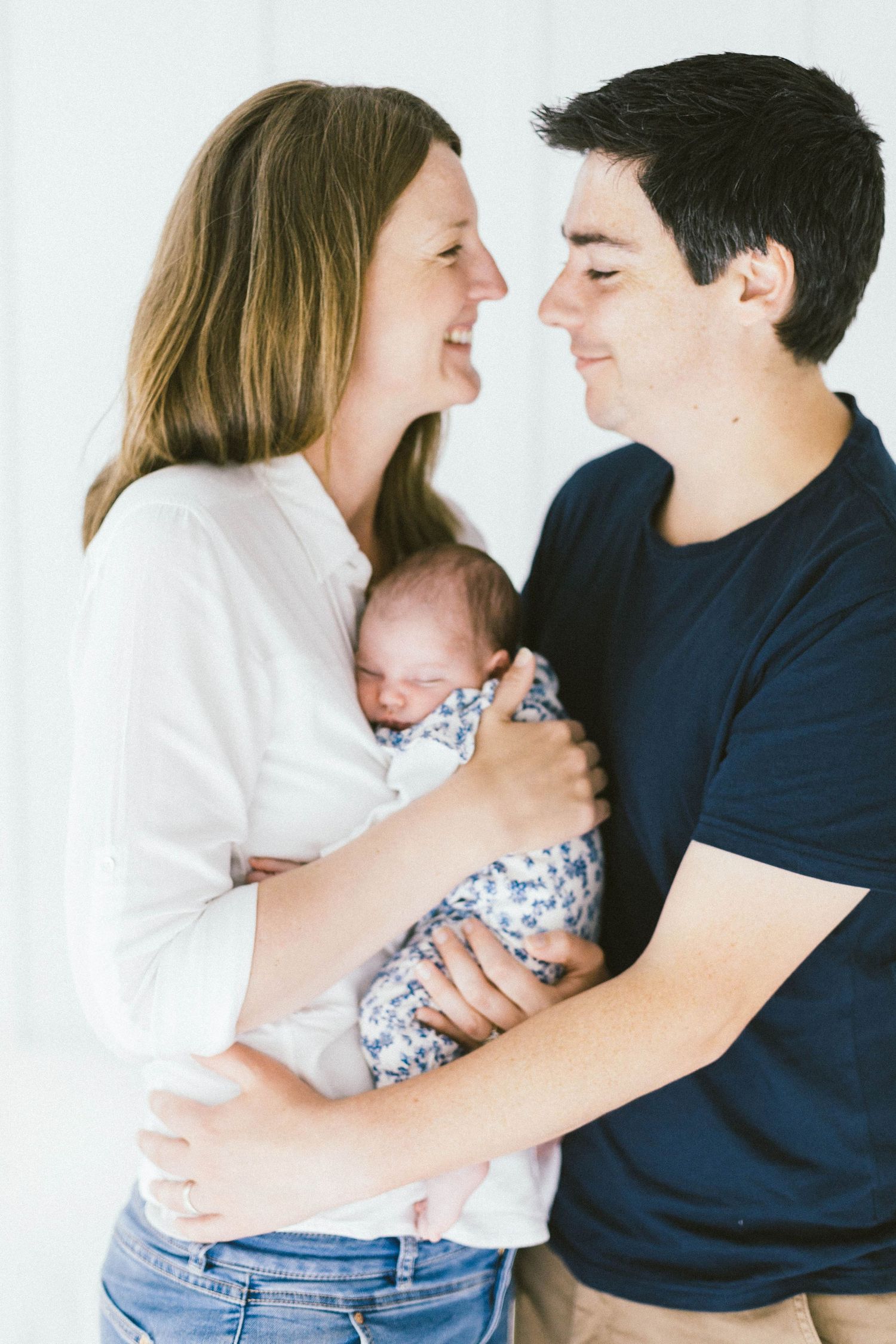 A couple shares an intimate moment while holding their newborn baby dressed in a floral outfit against a white backdrop.