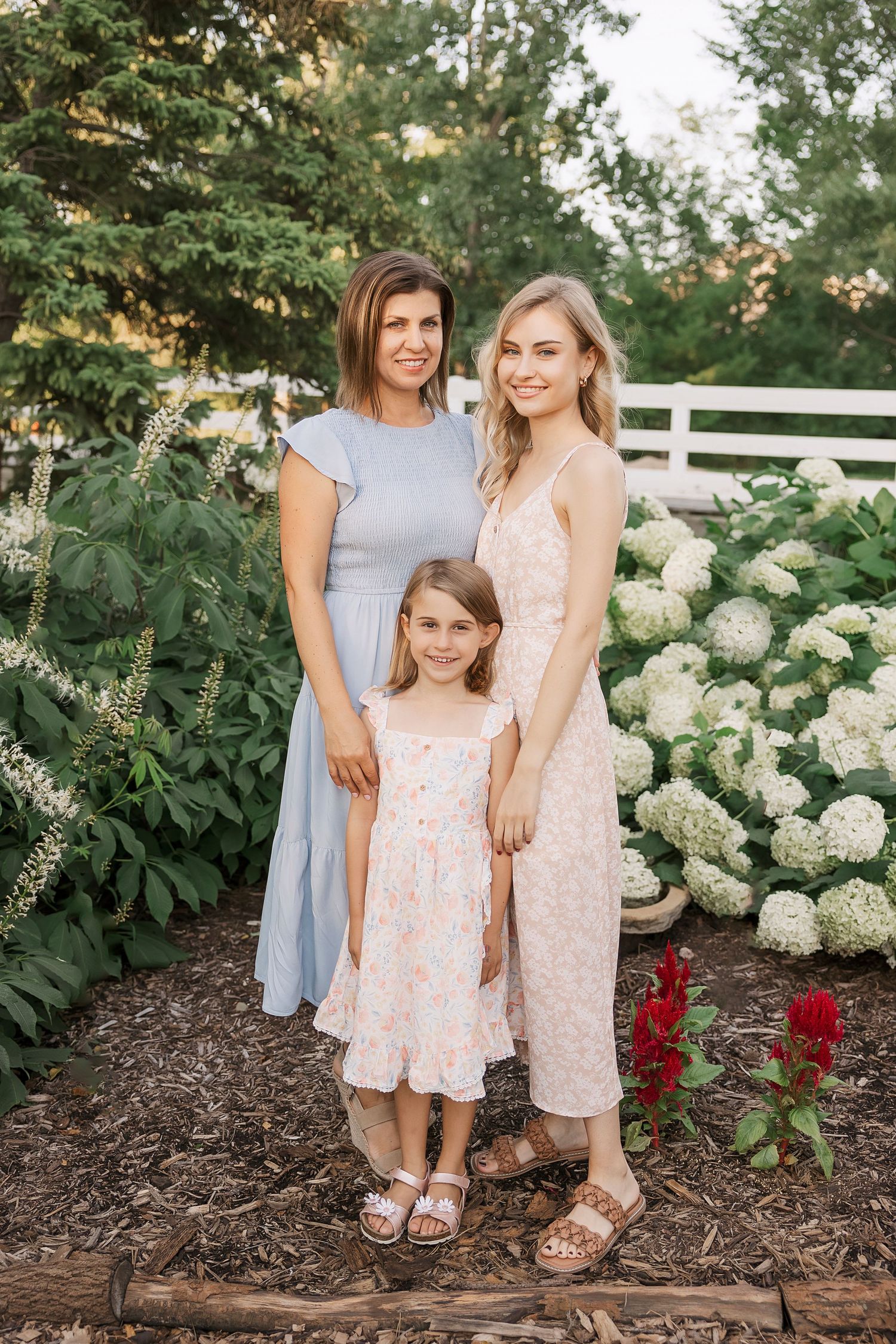 Three people pose together in garden with blooming white hydrangea bushes. Novi Northville Portrait Photographer