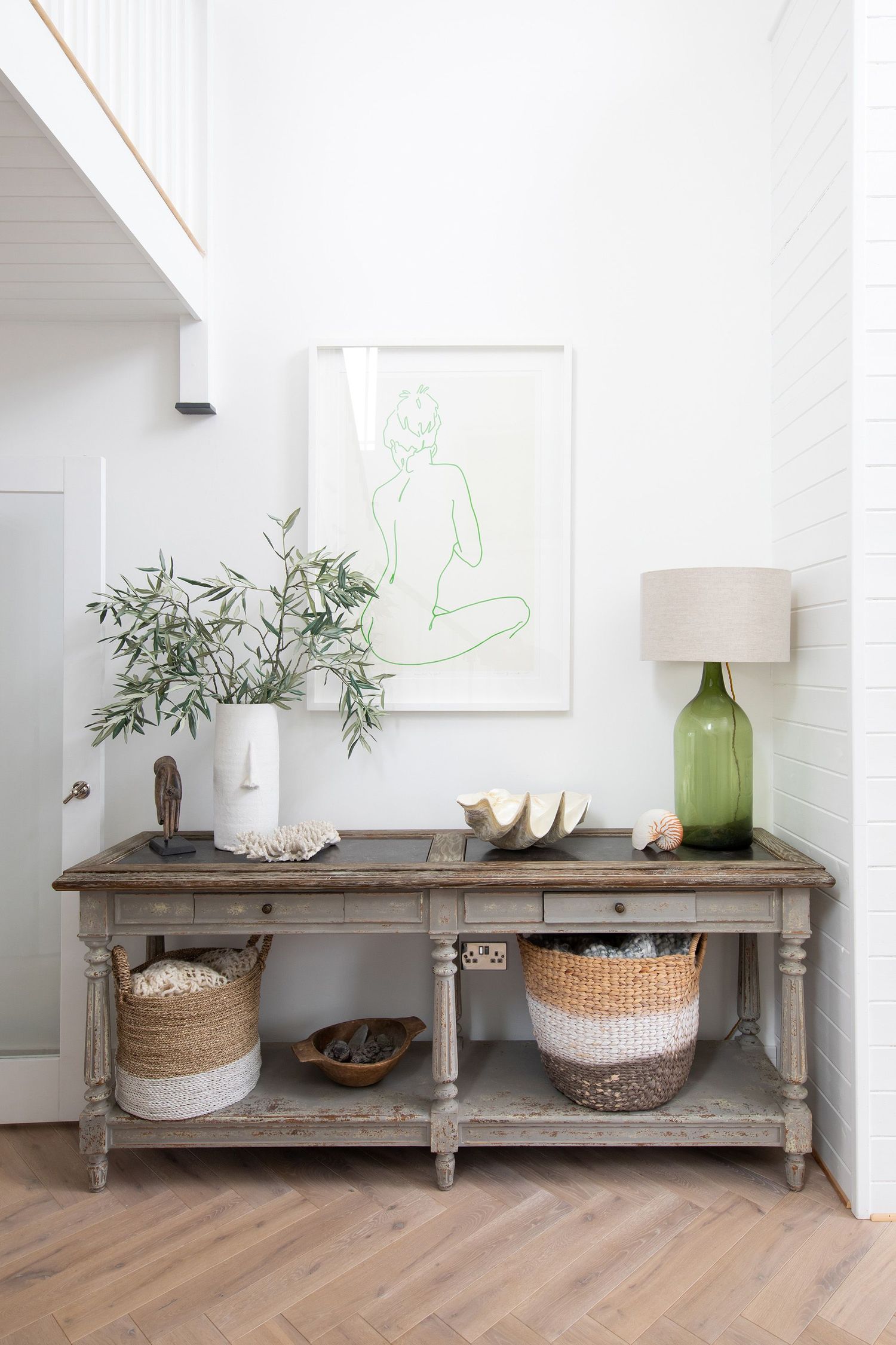Rustic console table with green lamp, minimalist art, and woven baskets beneath against white wall.