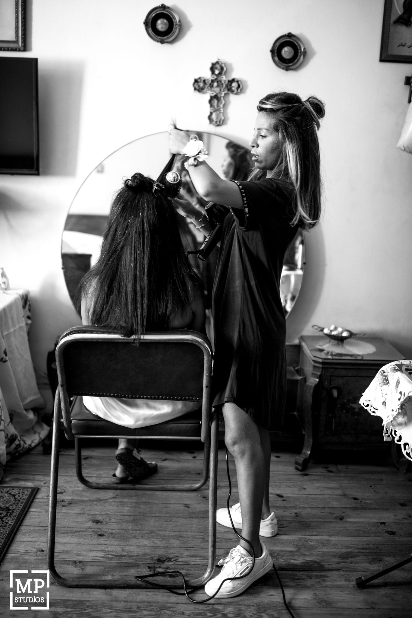 Black and white photo of styling hair in front of a round mirror with decorative crosses on wall.