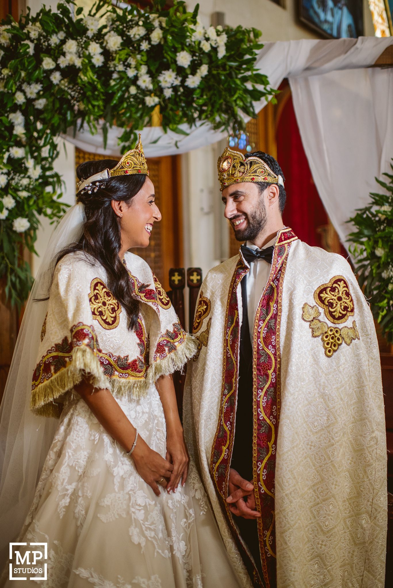 Couple wearing ornate traditional wedding crowns and embroidered ceremonial attire under a floral arch.