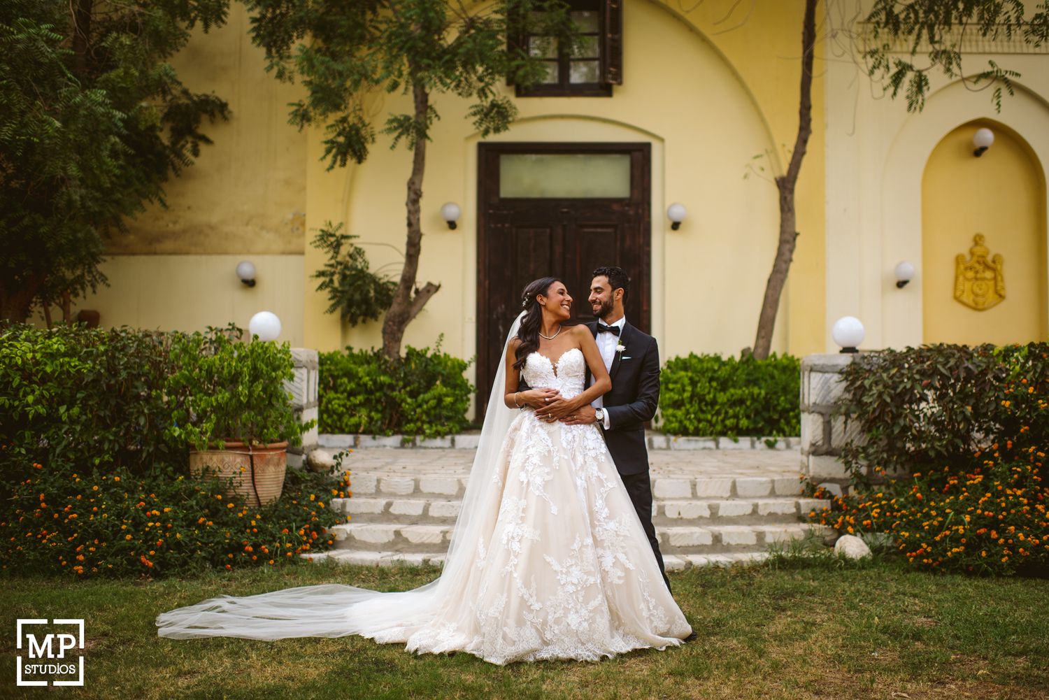 Two newly married people embrace on the steps of a yellow Spanish-style building with white lace dress and veil details.