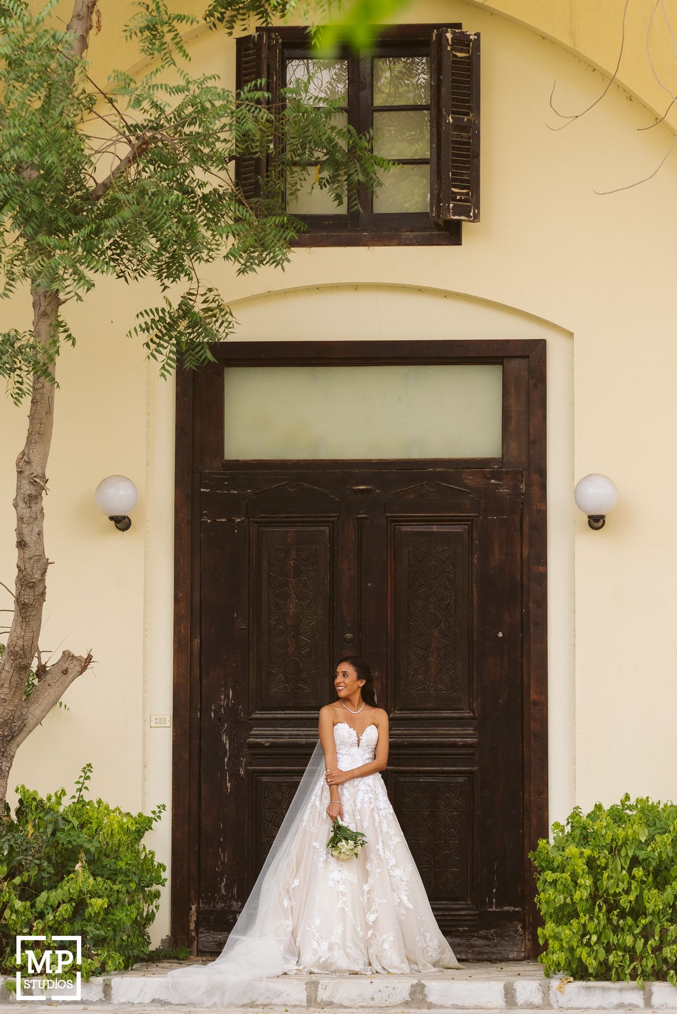 Beautiful white wedding dress photographed against a dark wooden door with yellow stucco walls and greenery.