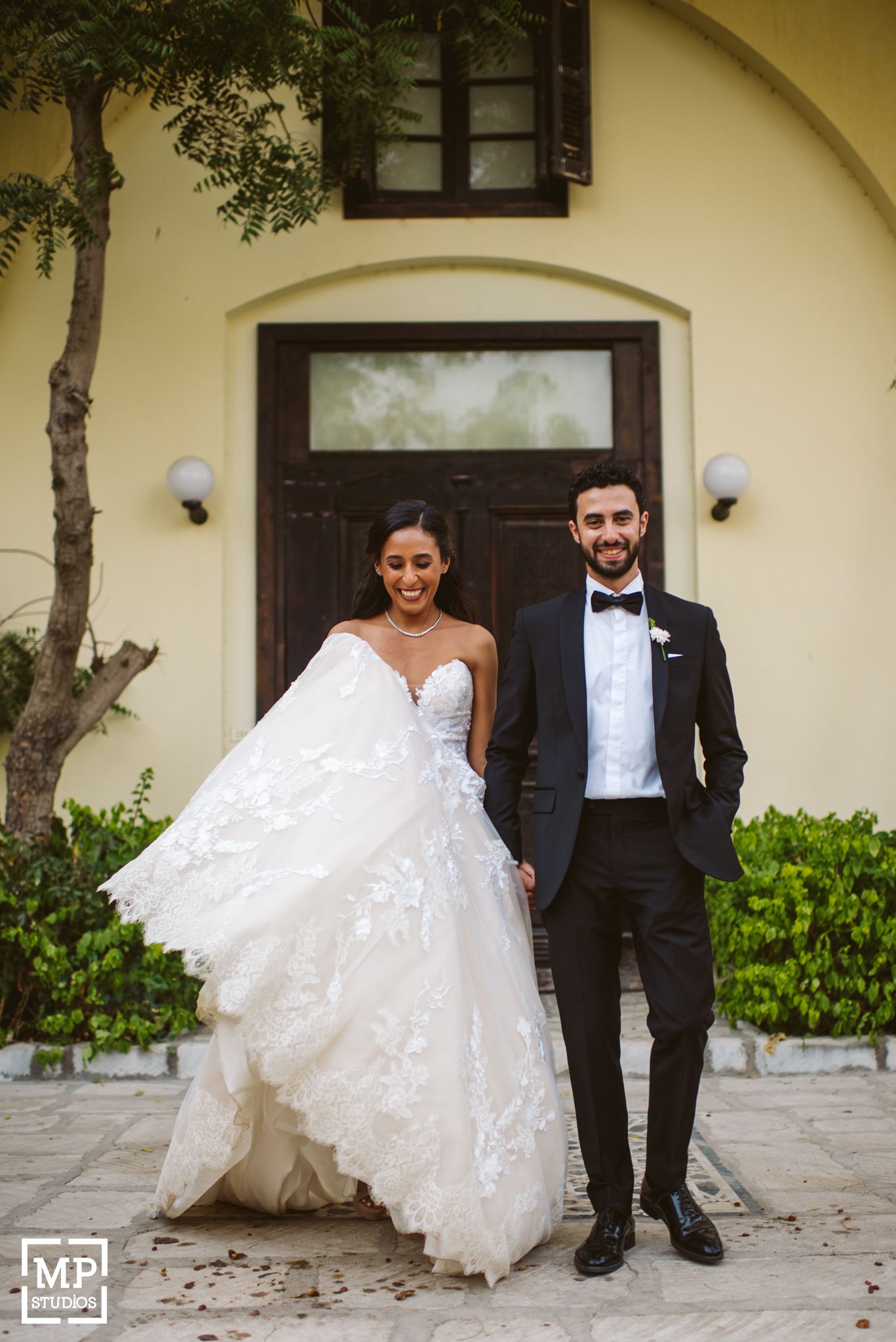 Elegant wedding portrait against yellow stucco wall and wooden door with flowing lace dress.