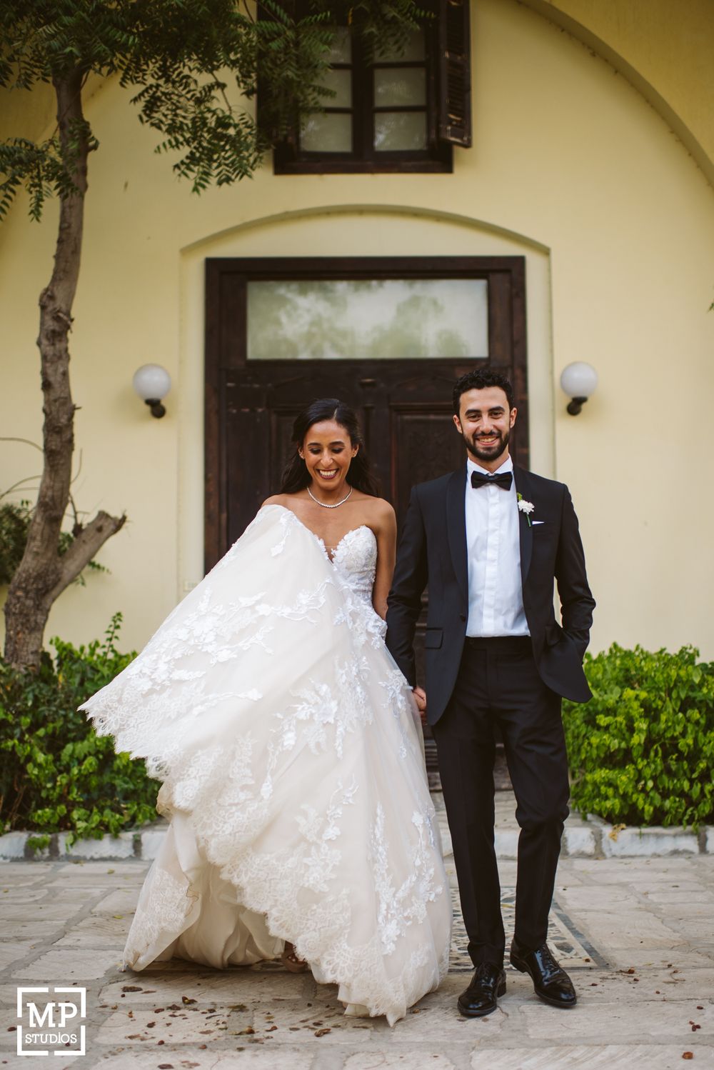Elegant wedding portrait against yellow stucco wall and wooden door with flowing lace dress.