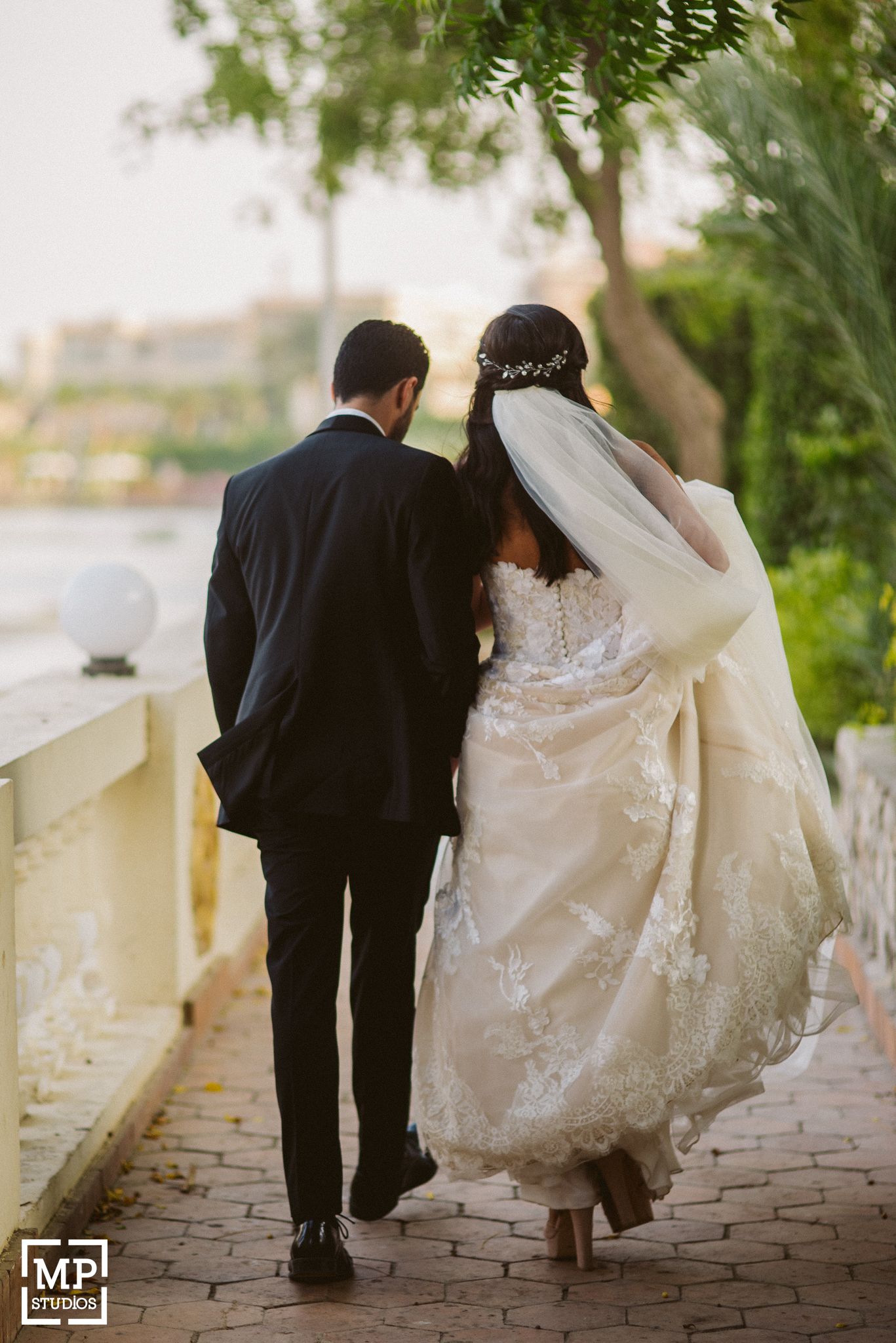 Romantic wedding photo from behind as couple walks along stone path surrounded by greenery.