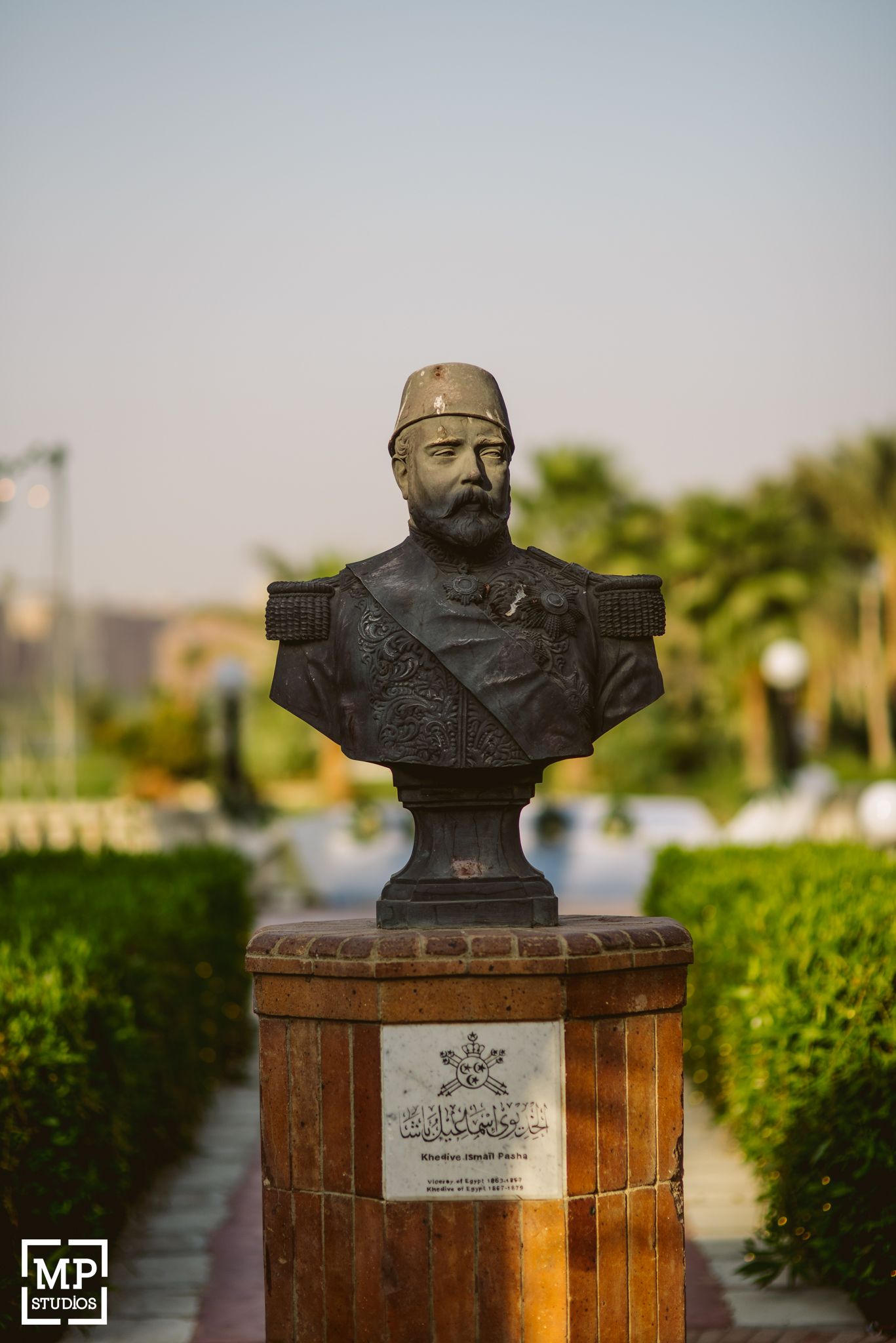 Bronze bust sculpture of a military officer with a beard and cap displayed on a wooden pedestal outdoors.