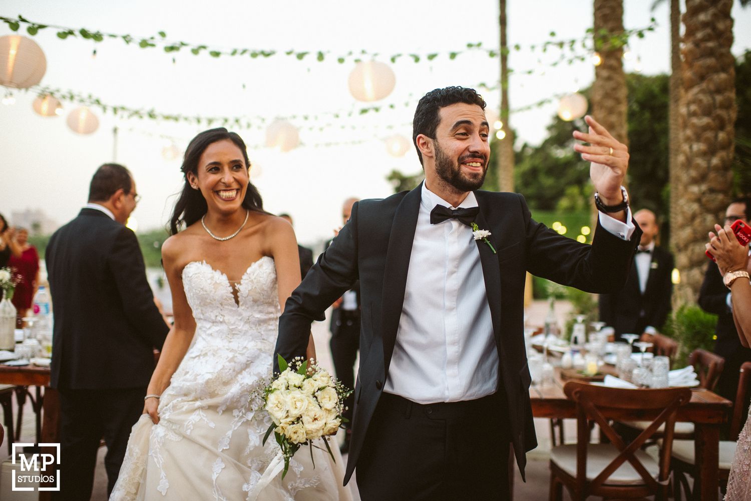 Elegant outdoor wedding celebration with hanging lights and palm trees in background.