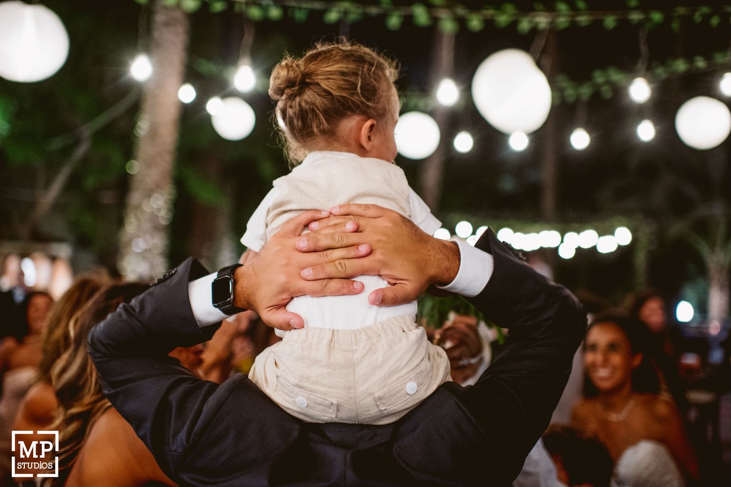 Parent holds small child wearing white formal attire during outdoor wedding reception.
