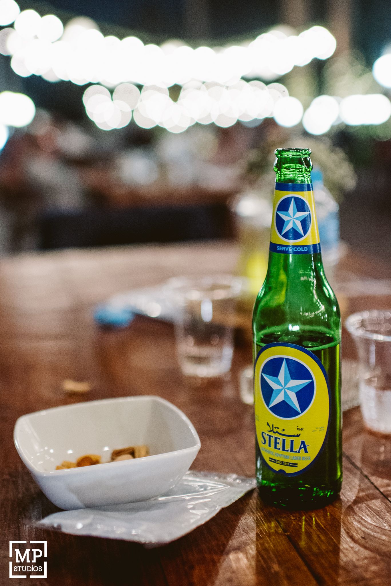A green bottle of Stella beer sits on a wooden table with bokeh lights in the background and a small dish nearby.