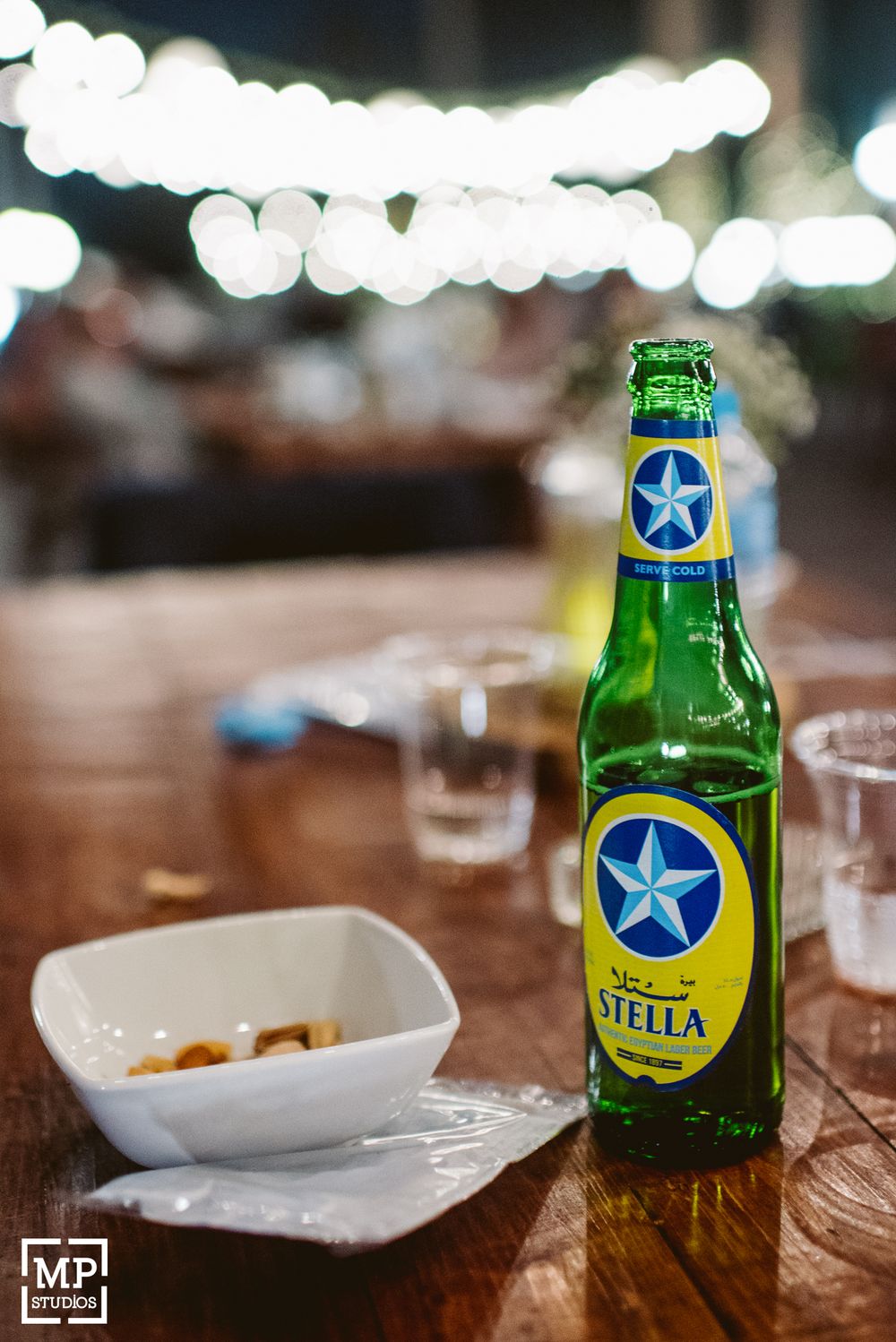 A green bottle of Stella beer sits on a wooden table with bokeh lights in the background and a small dish nearby.