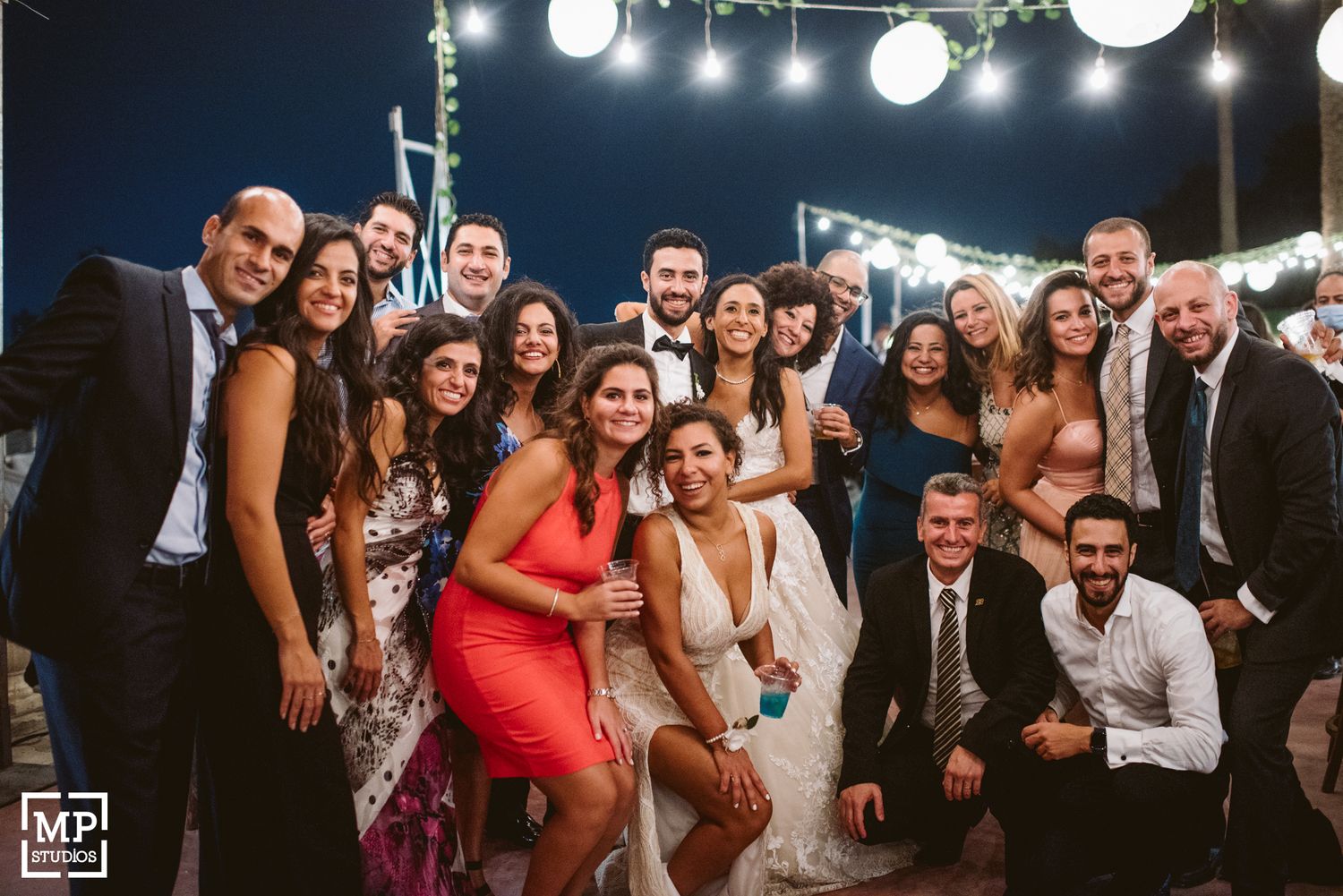 Large group photo of wedding guests posing together under evening string lights at an outdoor reception.