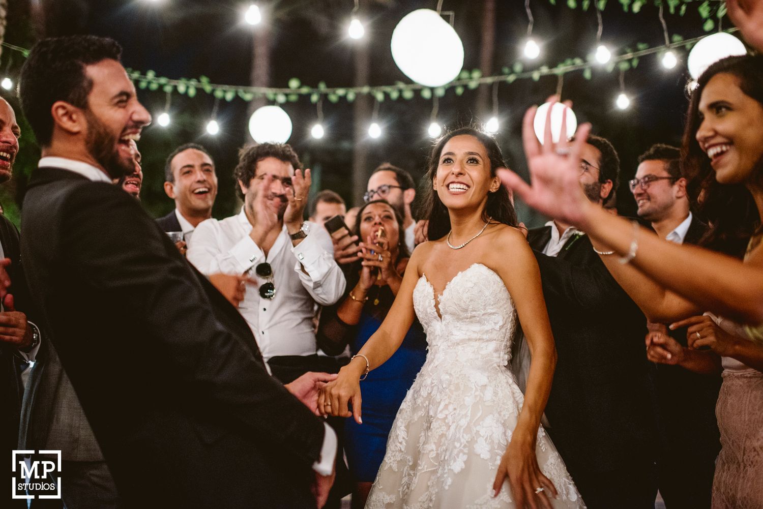 Wedding celebration on the dance floor with guests clapping and dancing under string lights.