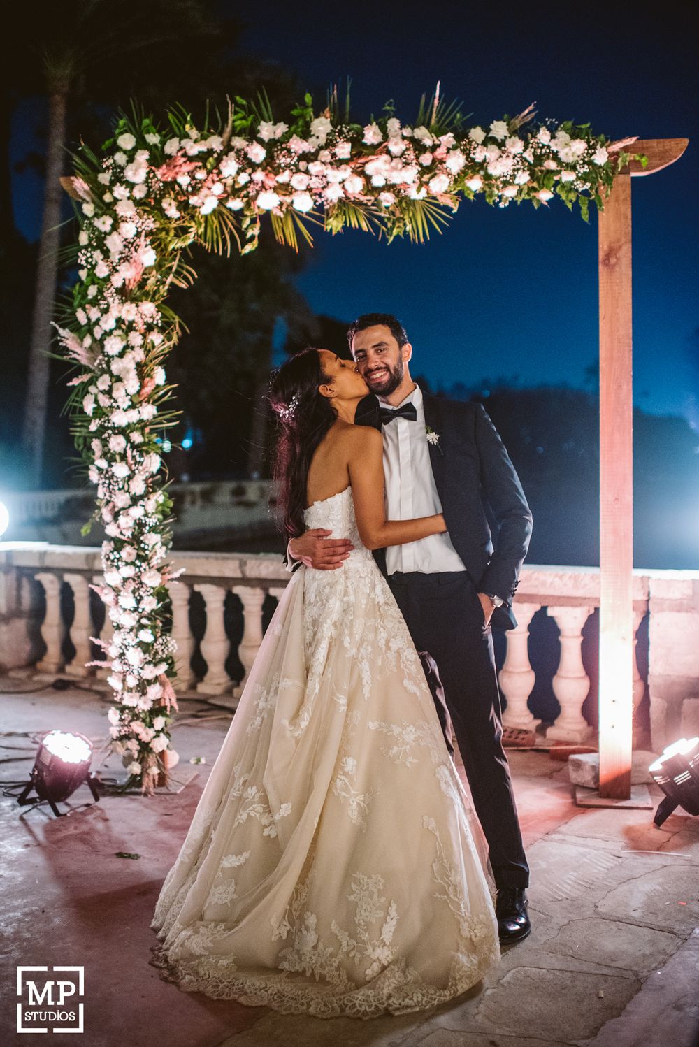 A romantic wedding photo under a floral arch decorated with white flowers at night.