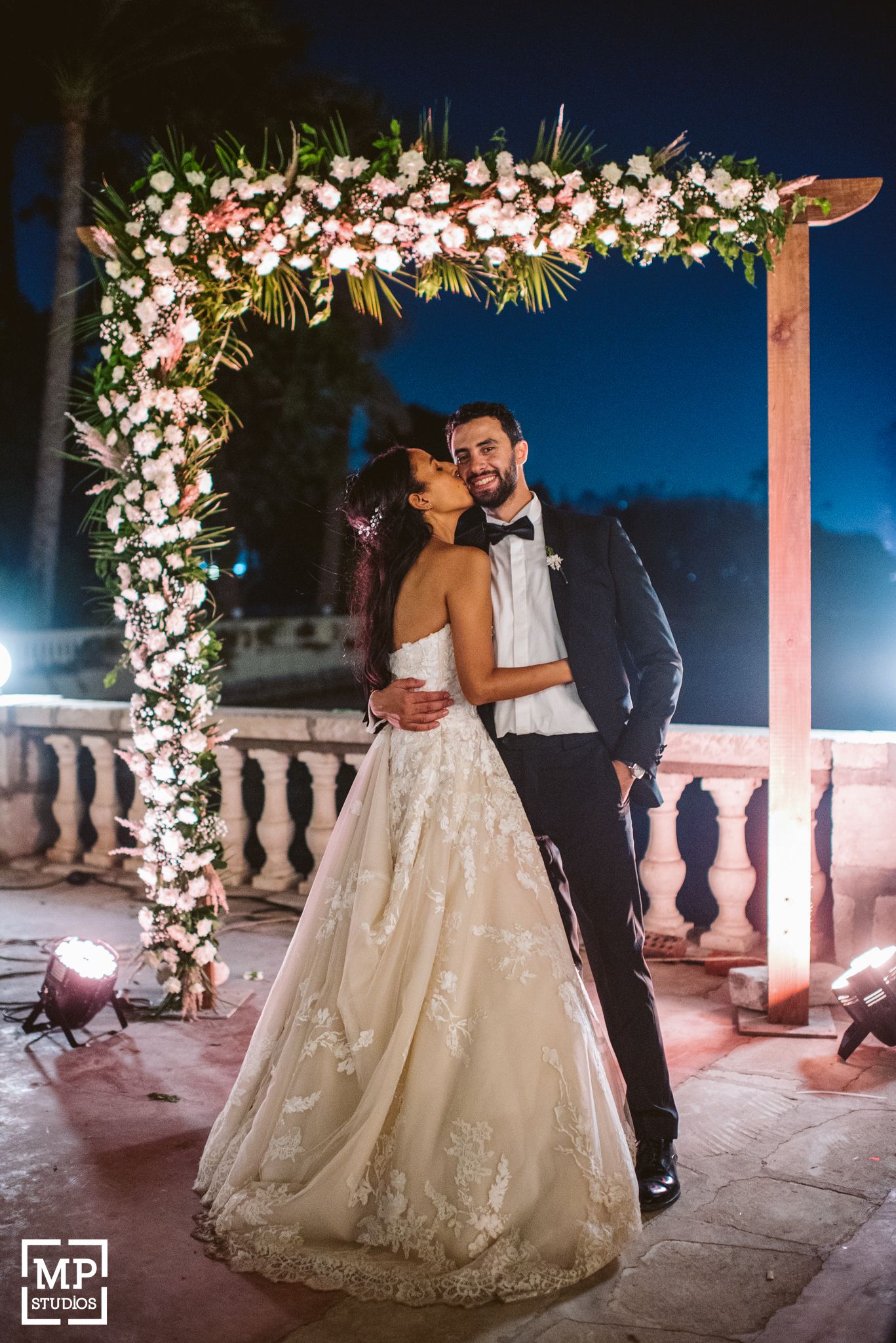 A romantic wedding photo under a floral arch decorated with white flowers at night.
