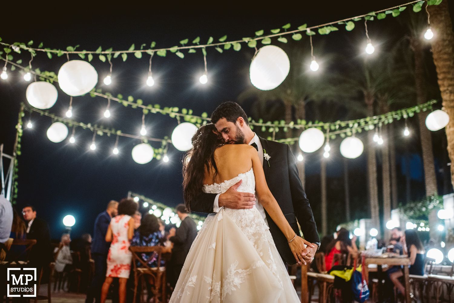 A romantic dance moment illuminated by hanging lanterns and string lights at an outdoor reception.